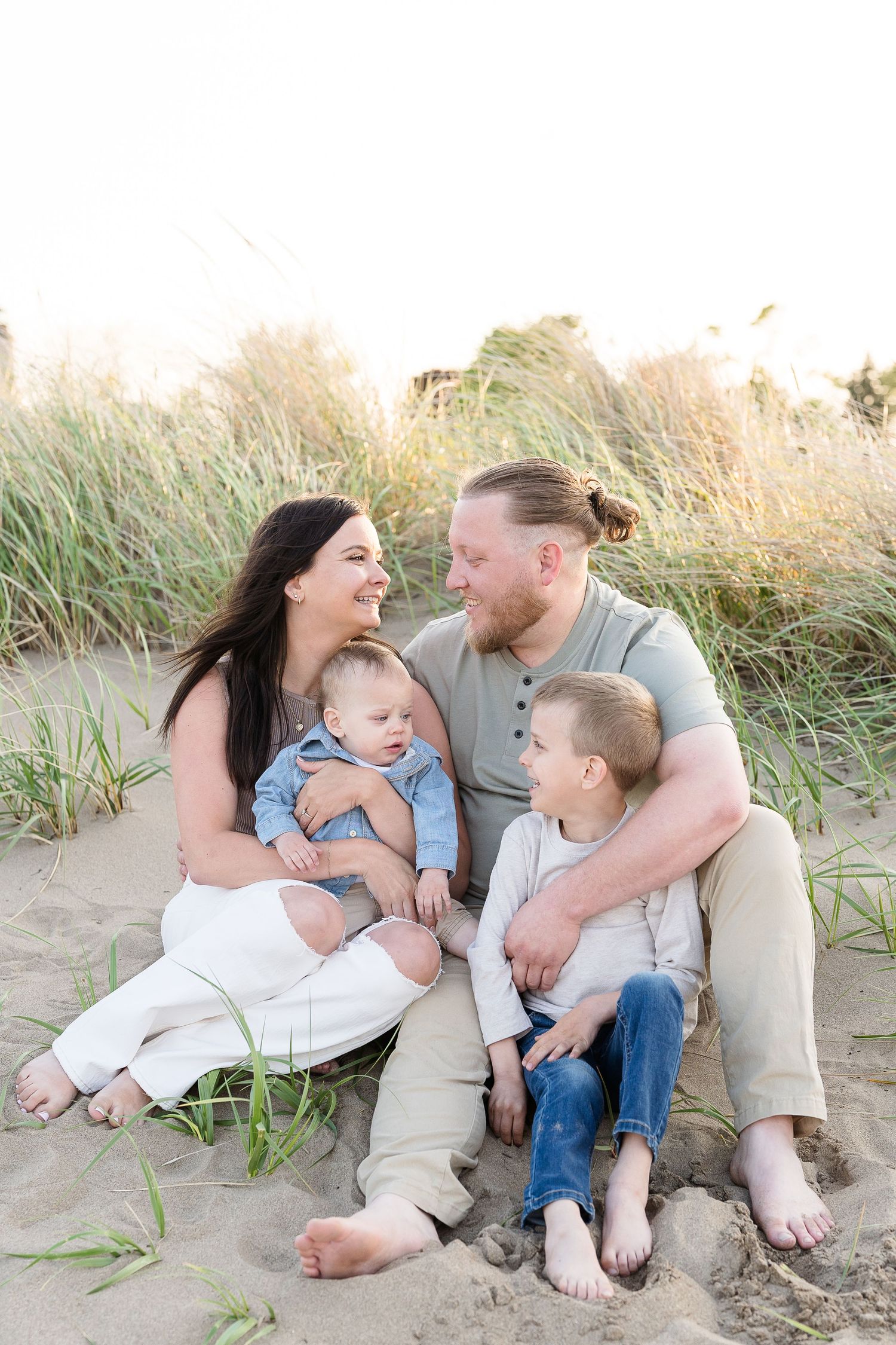 Loving family of four sitting barefoot on sandy beach dunes surrounded by tall grass in warm evening light.