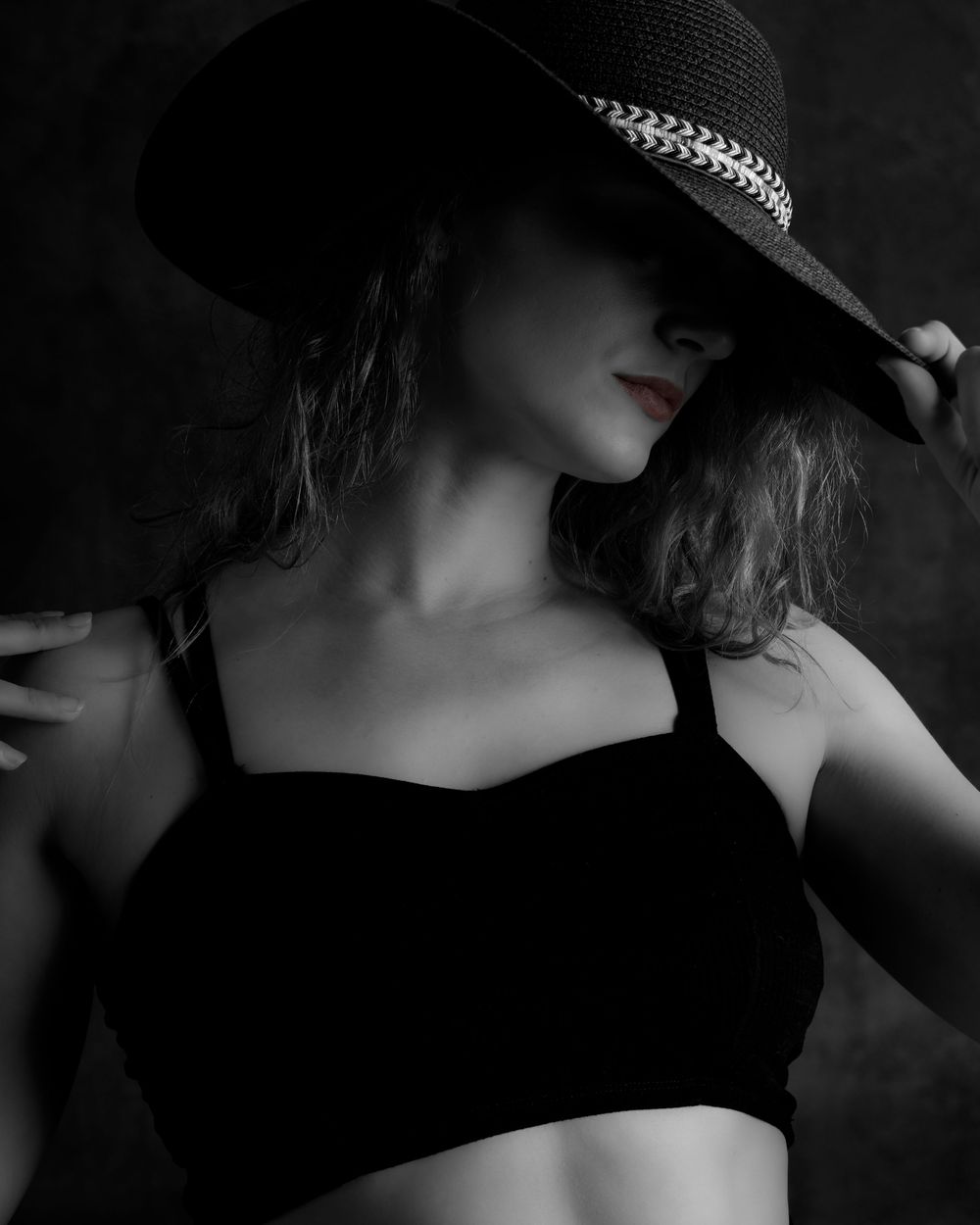 Black and white artistic portrait in film noir style showing a figure in a wide brimmed hat and black top against dark background.
