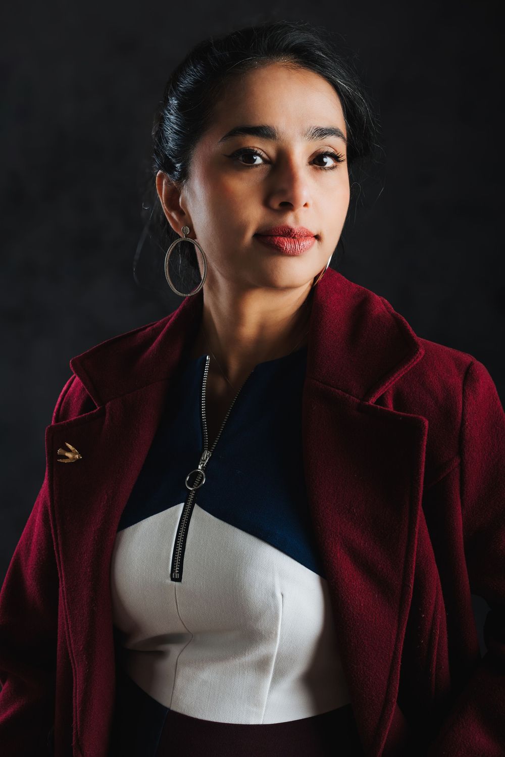 Studio portrait against dark background wearing a burgundy velvet jacket and white top with silver hoop earrings.