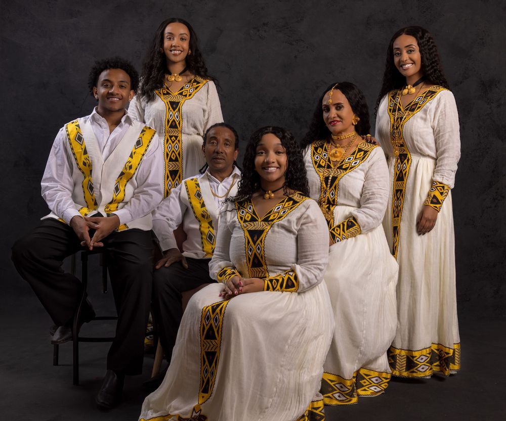 Five people wearing traditional Ethiopian formal attire with white fabric and gold embellishments pose together for a portrait.
