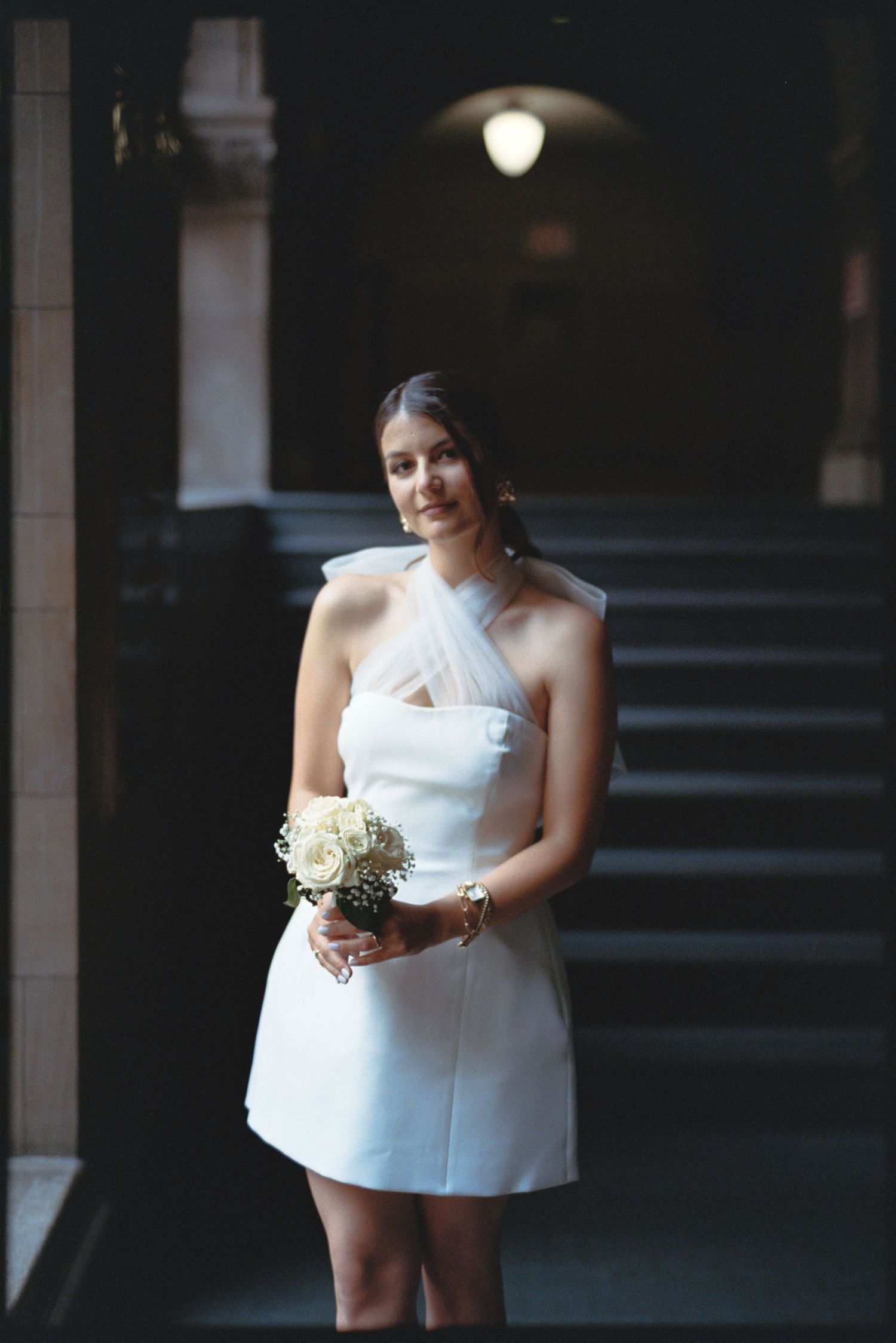 A bride in a short white dress holds a bouquet while standing in a dimly lit archway.