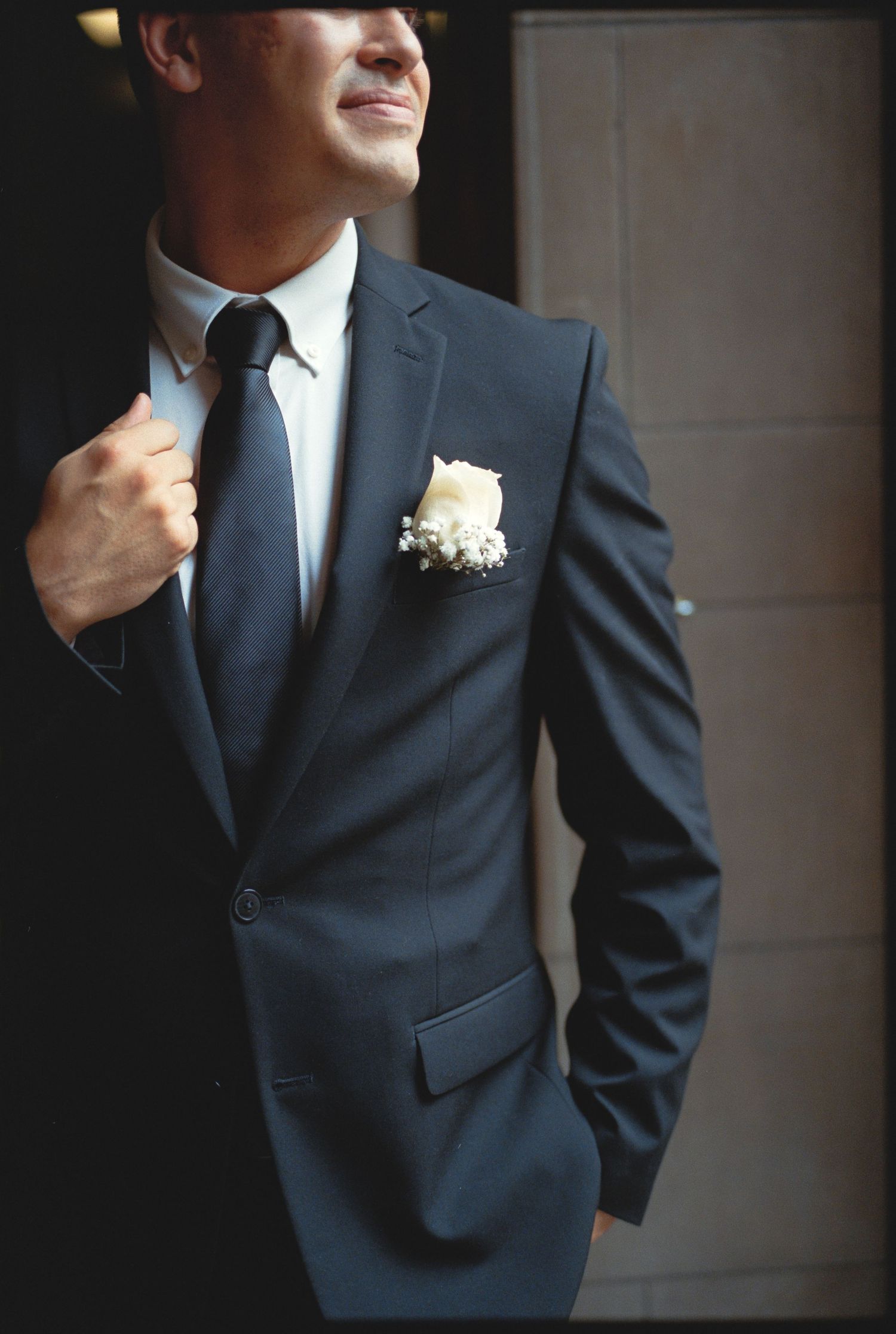 A groom in a navy suit adjusts his tie, with a white boutonniere on his lapel.