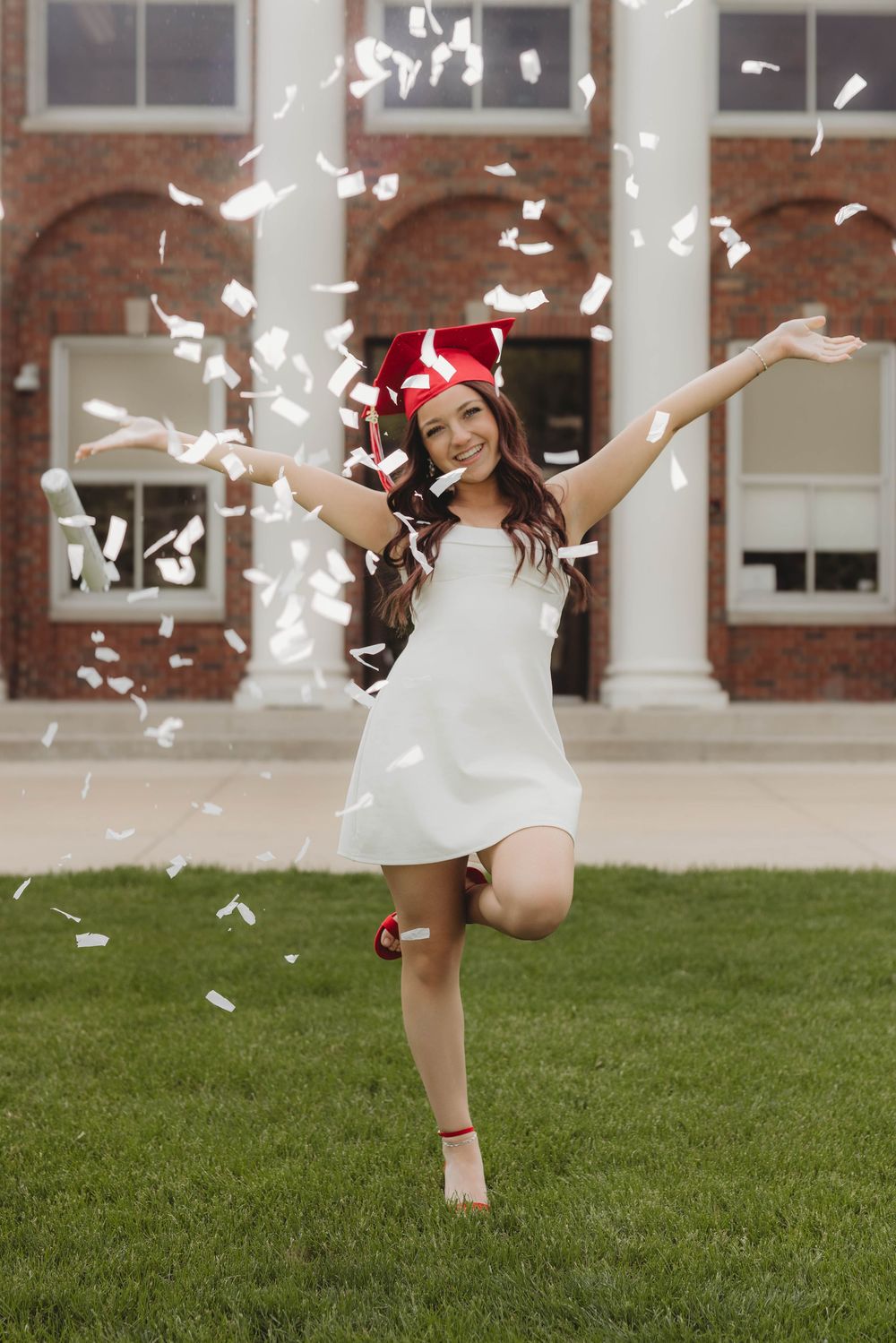 A joyful graduate in white dress and red cap celebrates with confetti flying around her outside a brick building.