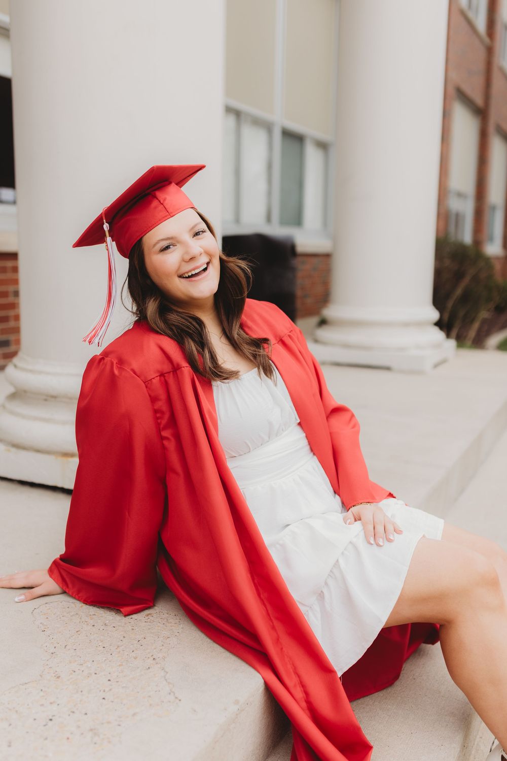 Graduate in red cap and gown poses playfully on concrete steps near brick building.