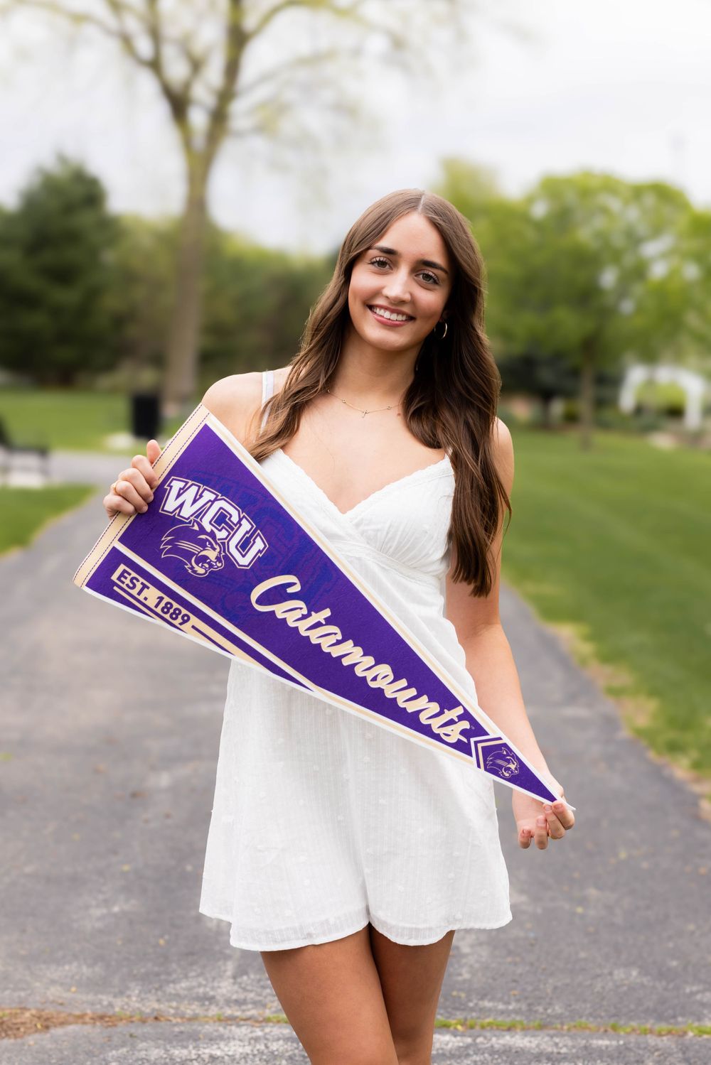 Young woman in white dress holding a purple WCU Catamounts pennant flag outdoors on a tree-lined path.