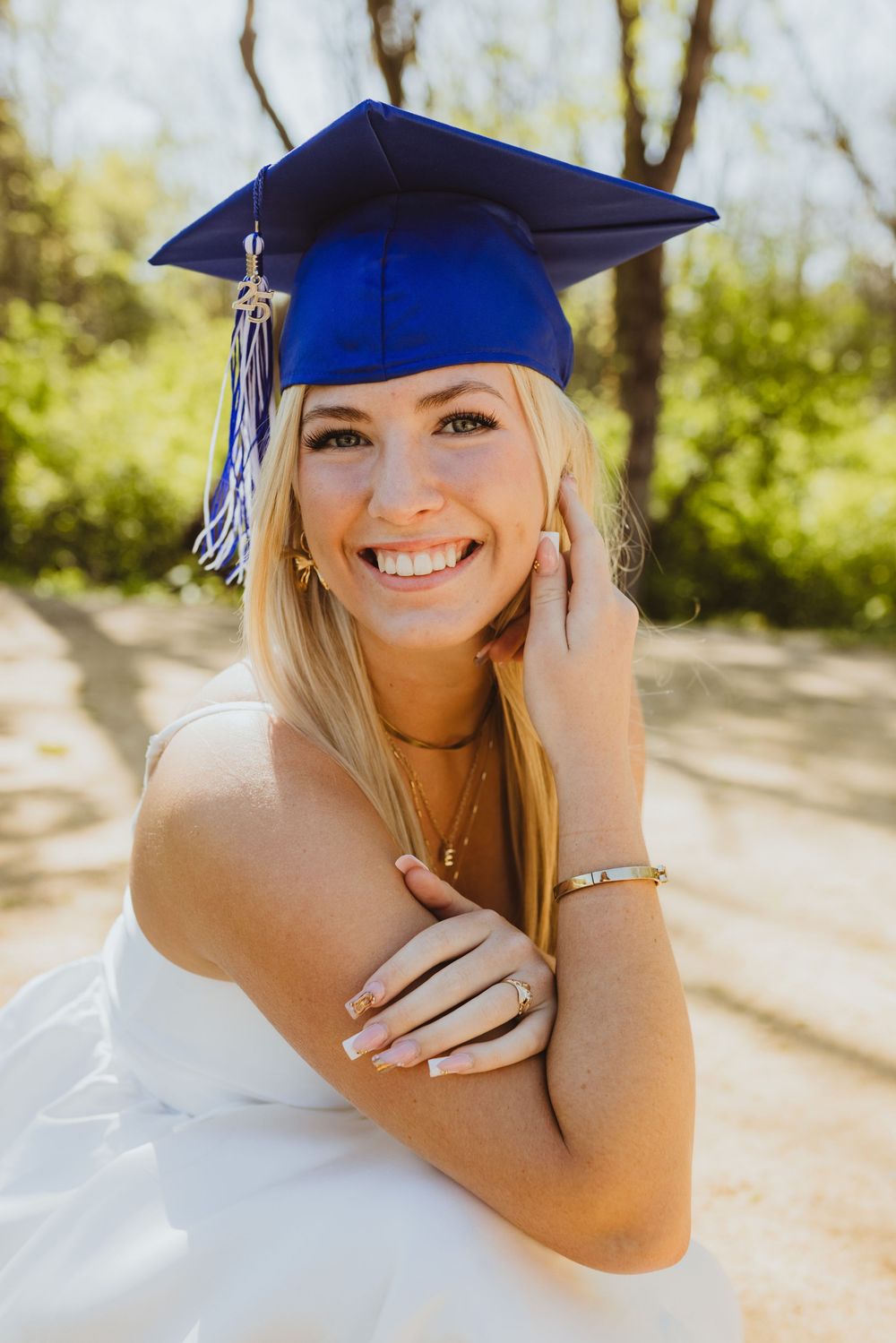 Graduate in blue cap and gown smiles brightly while wearing a white dress outdoors.