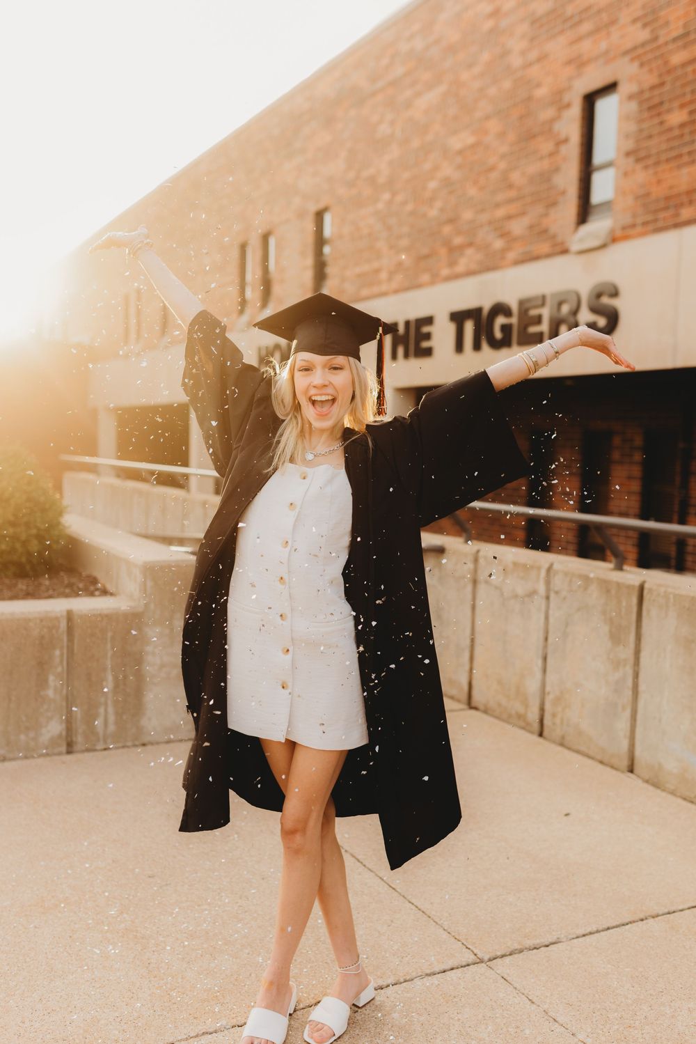 Graduate celebrates with glitter toss outside brick building during golden hour photoshoot.
