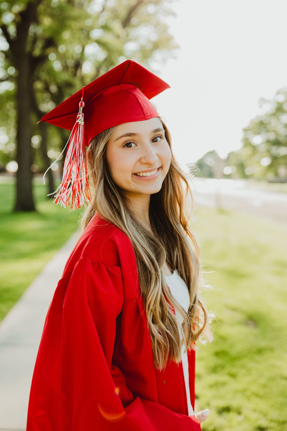 Graduate in red cap and gown posing outdoors on sunny grass field.