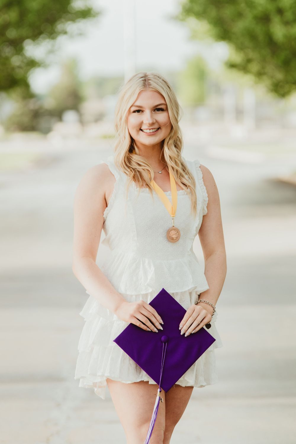 Graduate in white dress holding purple graduation cap outdoors.