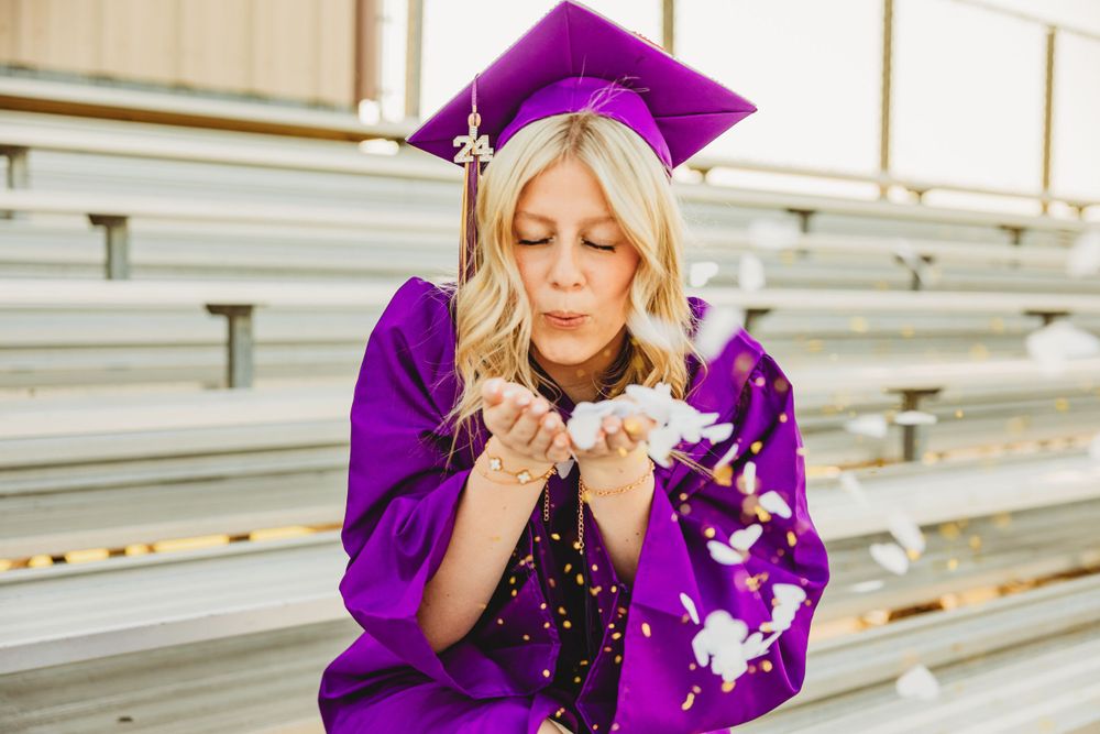 Graduate in purple cap and gown blows glitter confetti during outdoor graduation photo session.