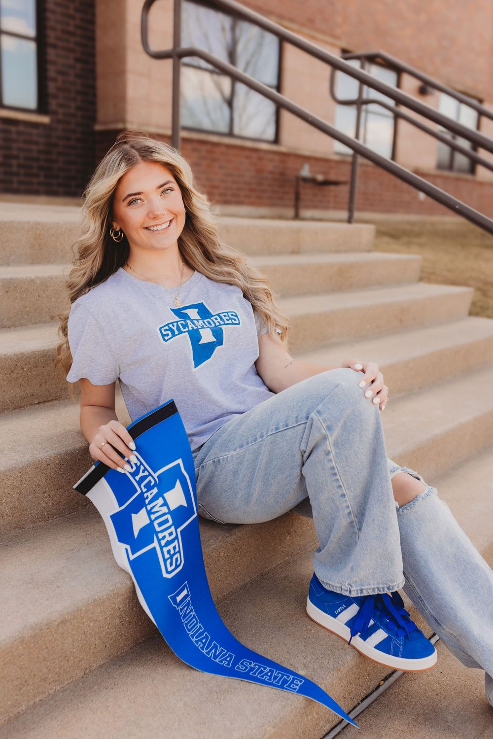 Graduate in casual attire sits on concrete steps holding blue graduation diploma cover.