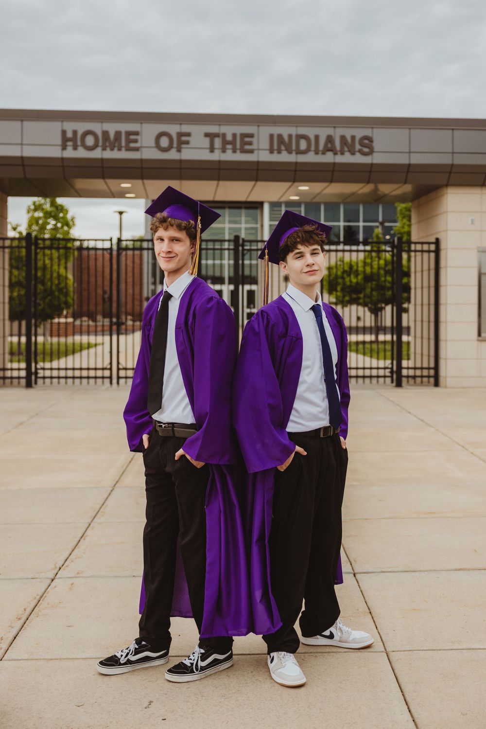 Two male graduates in purple caps and gowns pose back-to-back outside their school, Home of the Indians.