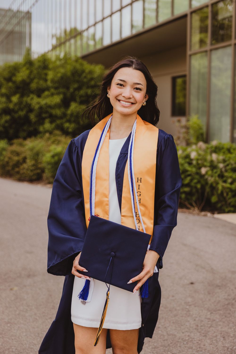 Smiling graduate in navy cap and gown with gold honor stole holds cap outdoors near modern building.