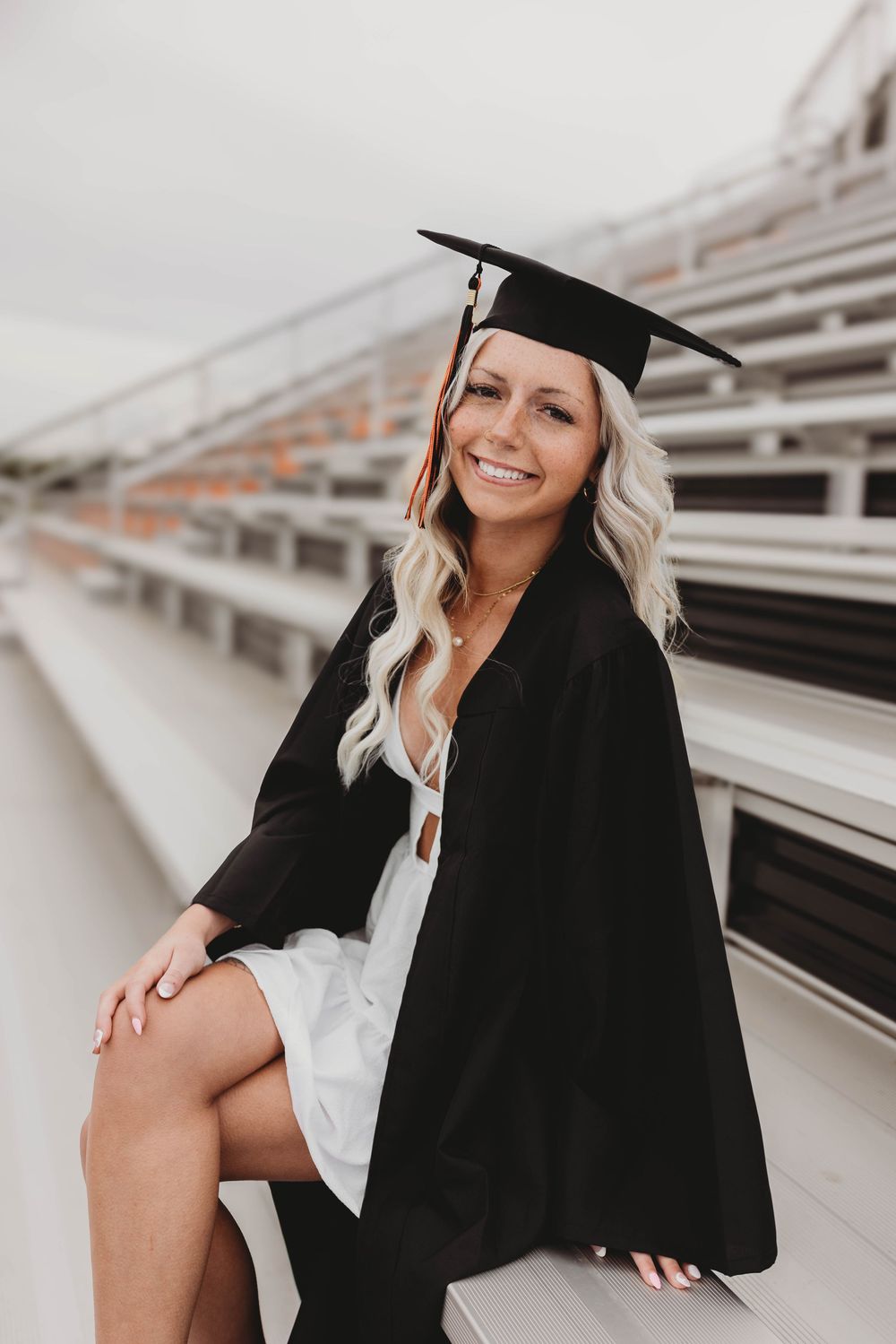 Smiling blonde woman in black graduation cap and gown sitting in stadium bleachers celebrating her graduation.