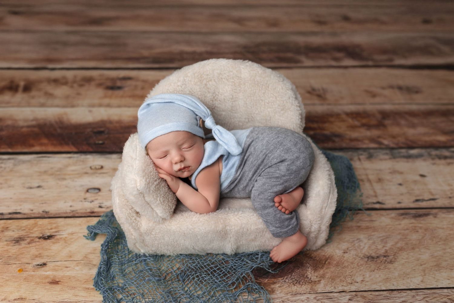 Sleeping newborn in white knit outfit on beige cushion against wooden floor.