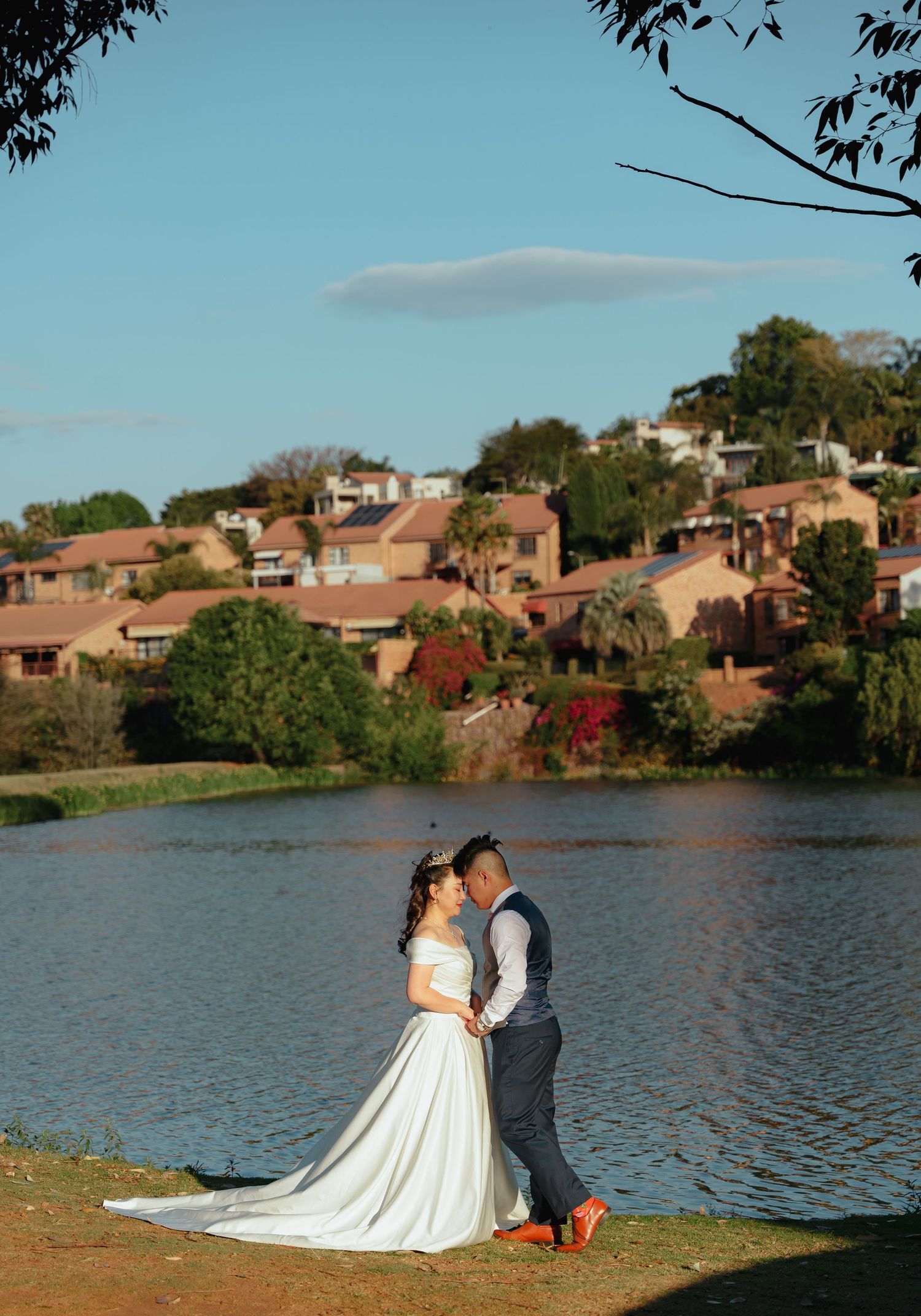Couple in wedding attire embracing by a lake with hillside houses in the background.