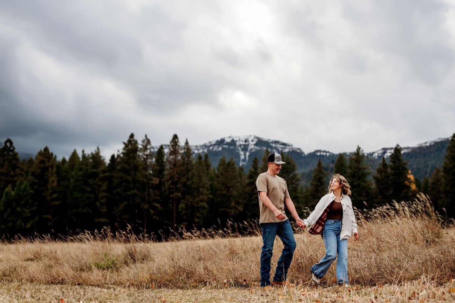 Couple walking hand in hand through autumn field with snow-capped mountains in distance. Couple Portrait Enterprise, Oregon