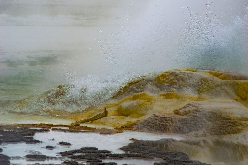 Yellow and brown mineral formations with steam and water droplets at a geothermal feature.