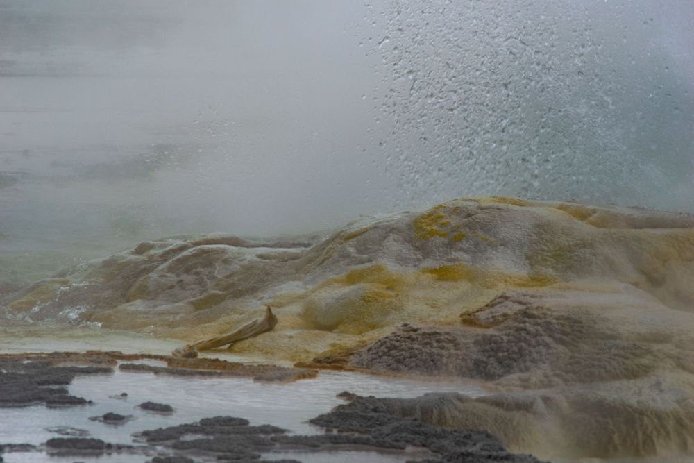 Steamy landscape with yellow and brown mineral deposits forming terraces near a hot spring.