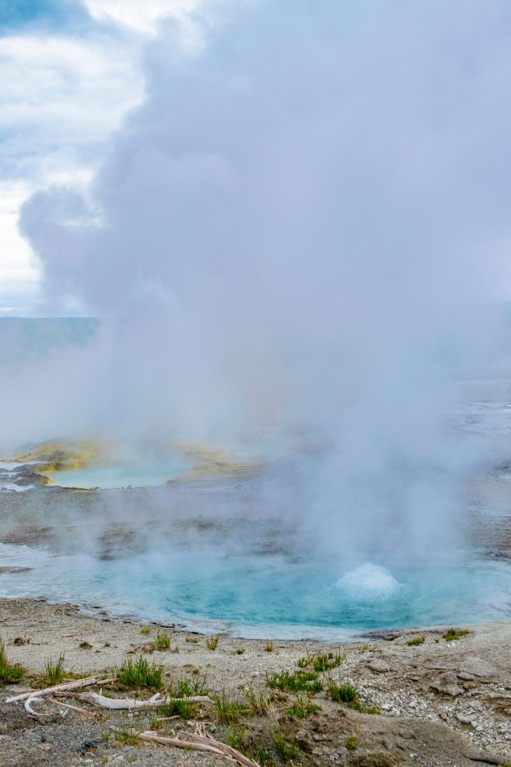 Turquoise hot spring emits steam under cloudy skies in a geothermal basin.