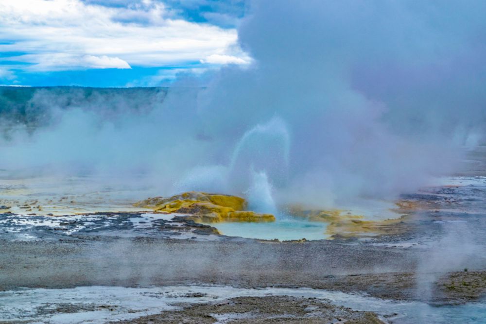 Vibrant blue and yellow hot spring with steam rising against a cloudy sky.