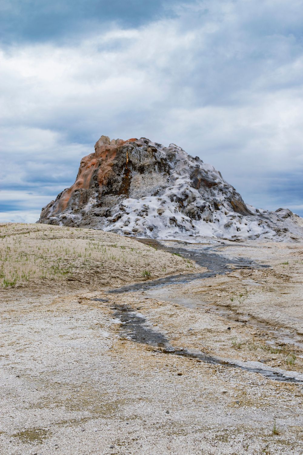 White and orange mineral deposits form a conical mound with steam venting in a barren landscape.