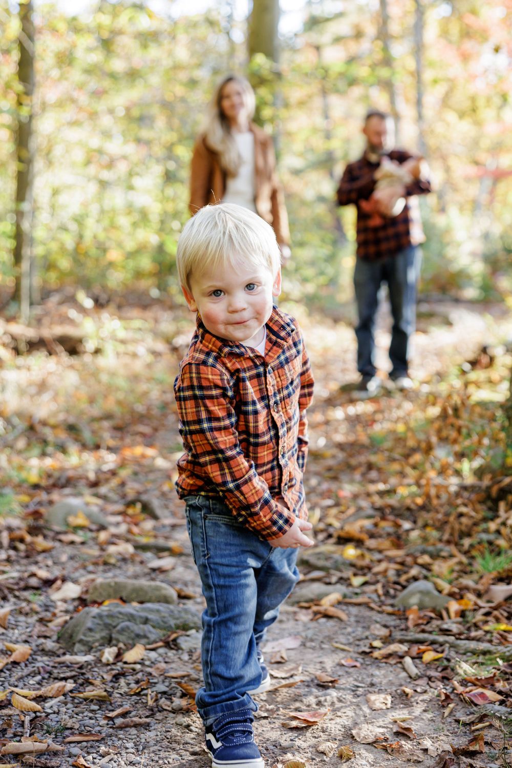 A child in a plaid flannel shirt and jeans plays in autumn leaves on a forest trail.