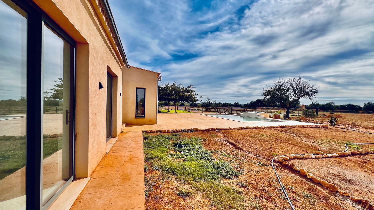 Modern stucco home exterior with large glass doors overlooking an undeveloped dirt yard under dramatic blue skies.