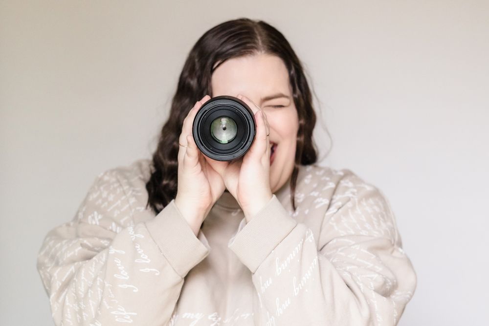 A photographer in a cozy sweater looks through a camera lens against a light background.