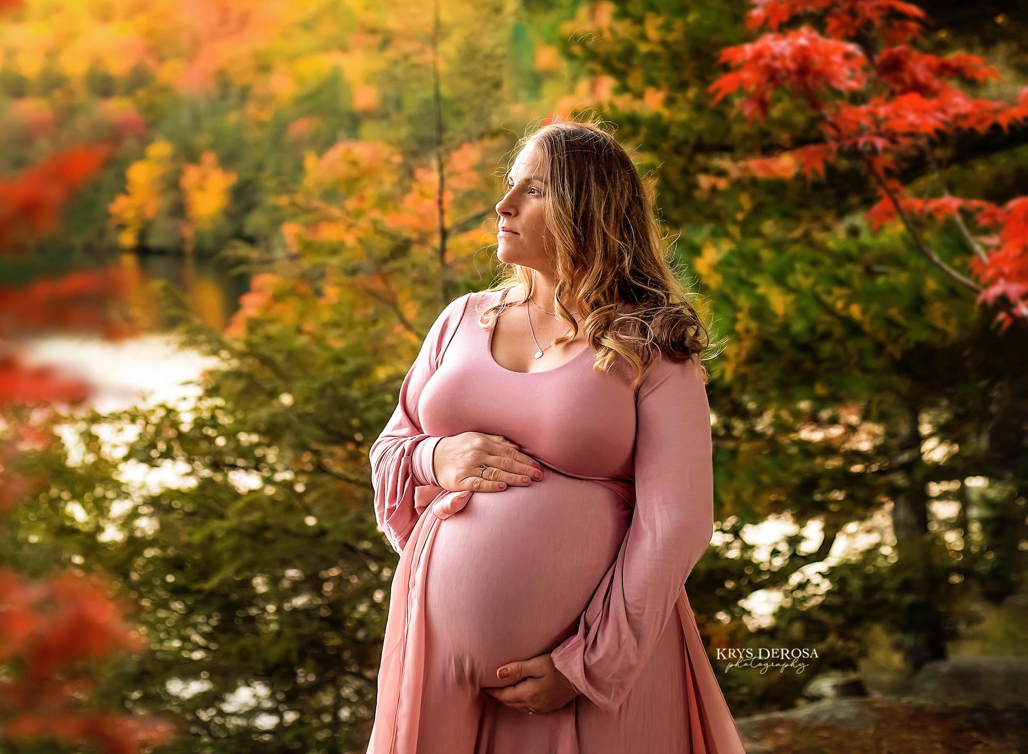 Expecting mother in a pink flowy dress poses in an autumn forest setting with vibrant red and orange fall foliage.