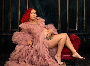 A glamorous portrait against a dark background features someone in a flowing pink ruffled gown reclining on red velvet furniture.