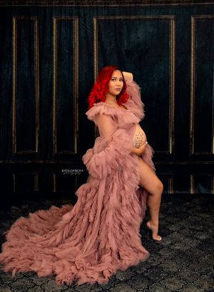 Pregnant woman in flowing pink tulle maternity gown with red hair poses elegantly against dark ornate backdrop.