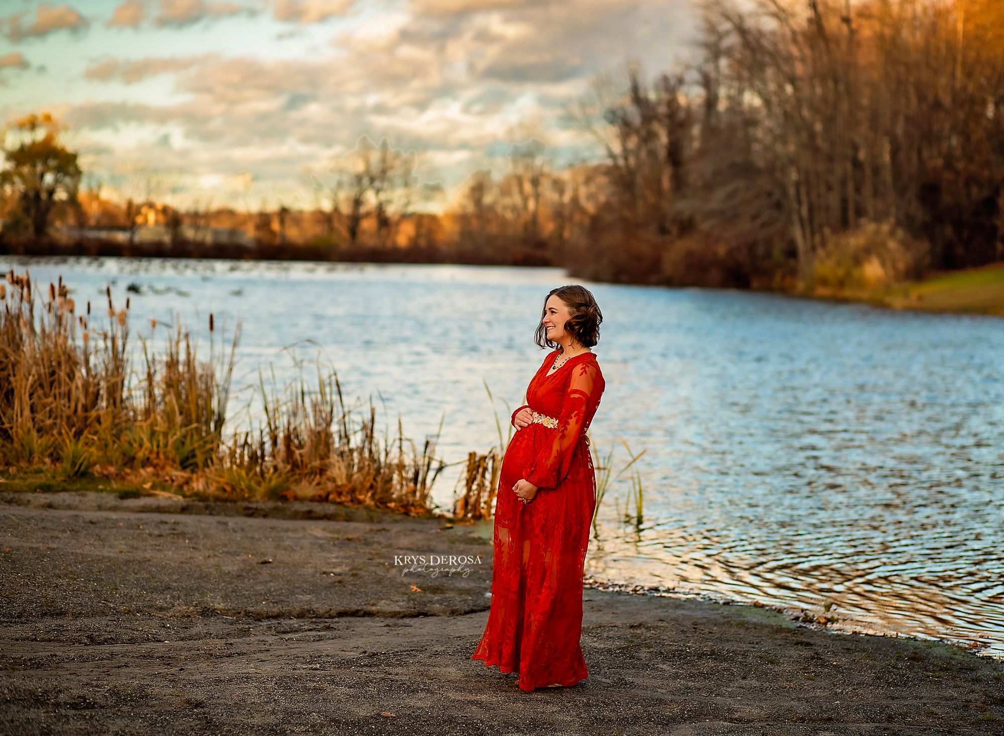 A person in a flowing red dress stands by a tranquil lake at sunset with autumn foliage in the background.