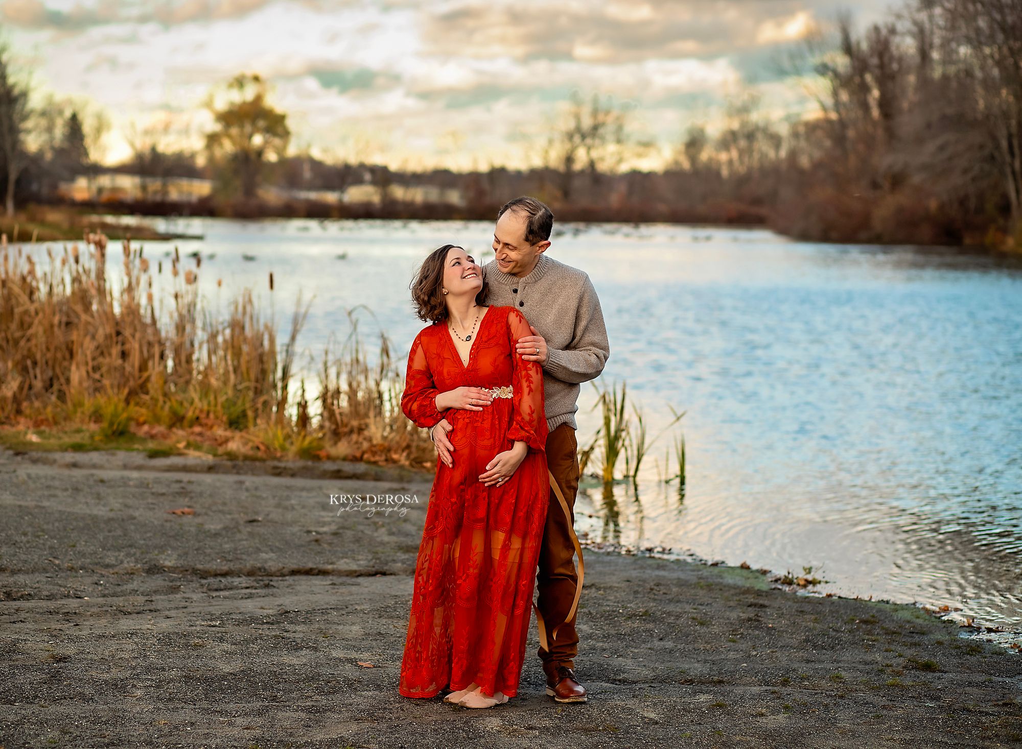 Couple maternity shoot outdoors by a lake, woman in red dress holding baby bump at sunset.
