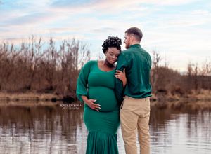 Pregnant couple in matching green outfits posing near a lake during a maternity photography session at sunset.