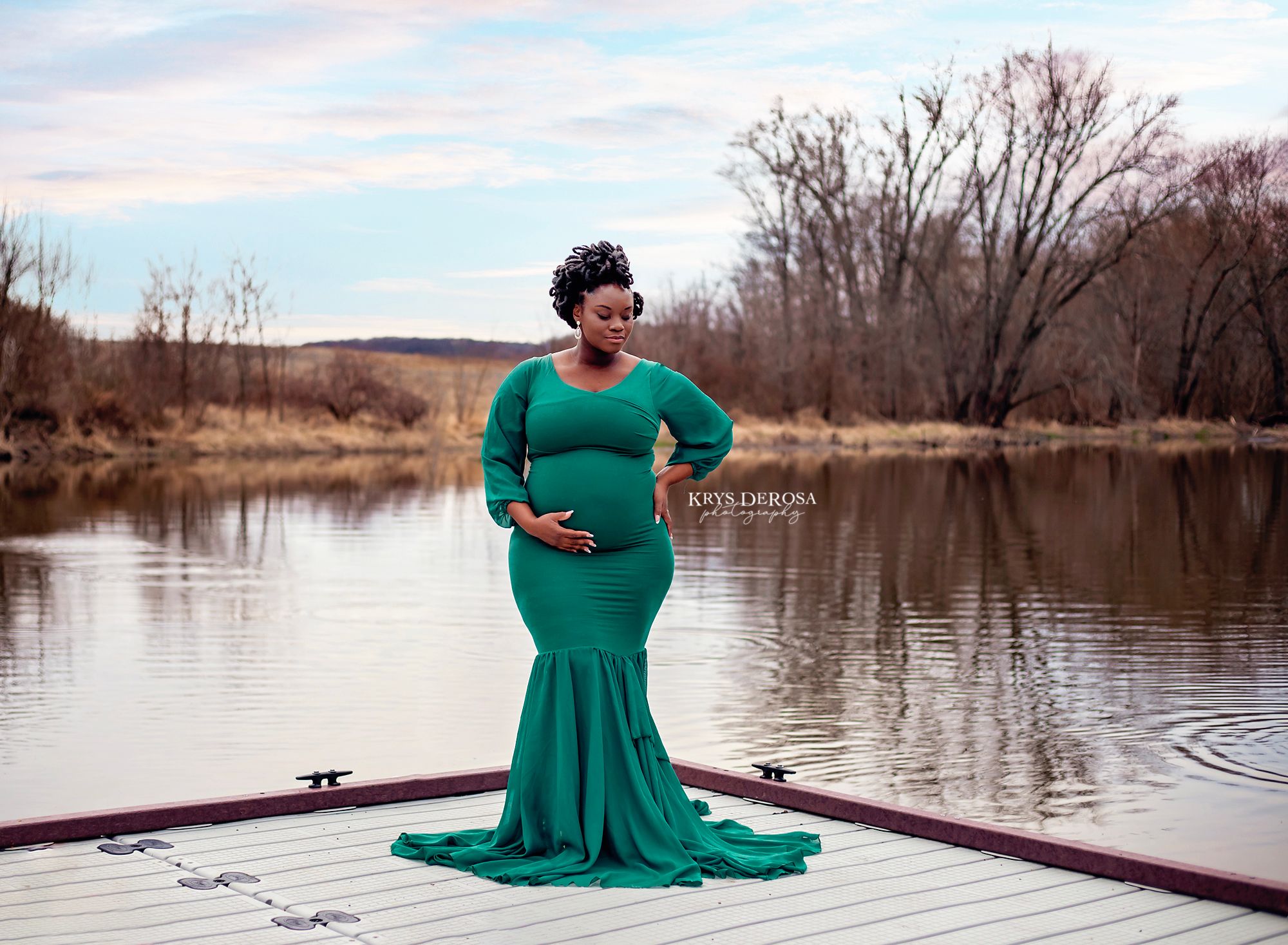 Pregnant woman in elegant green mermaid gown poses on a dock by a serene lake during an outdoor maternity photoshoot.