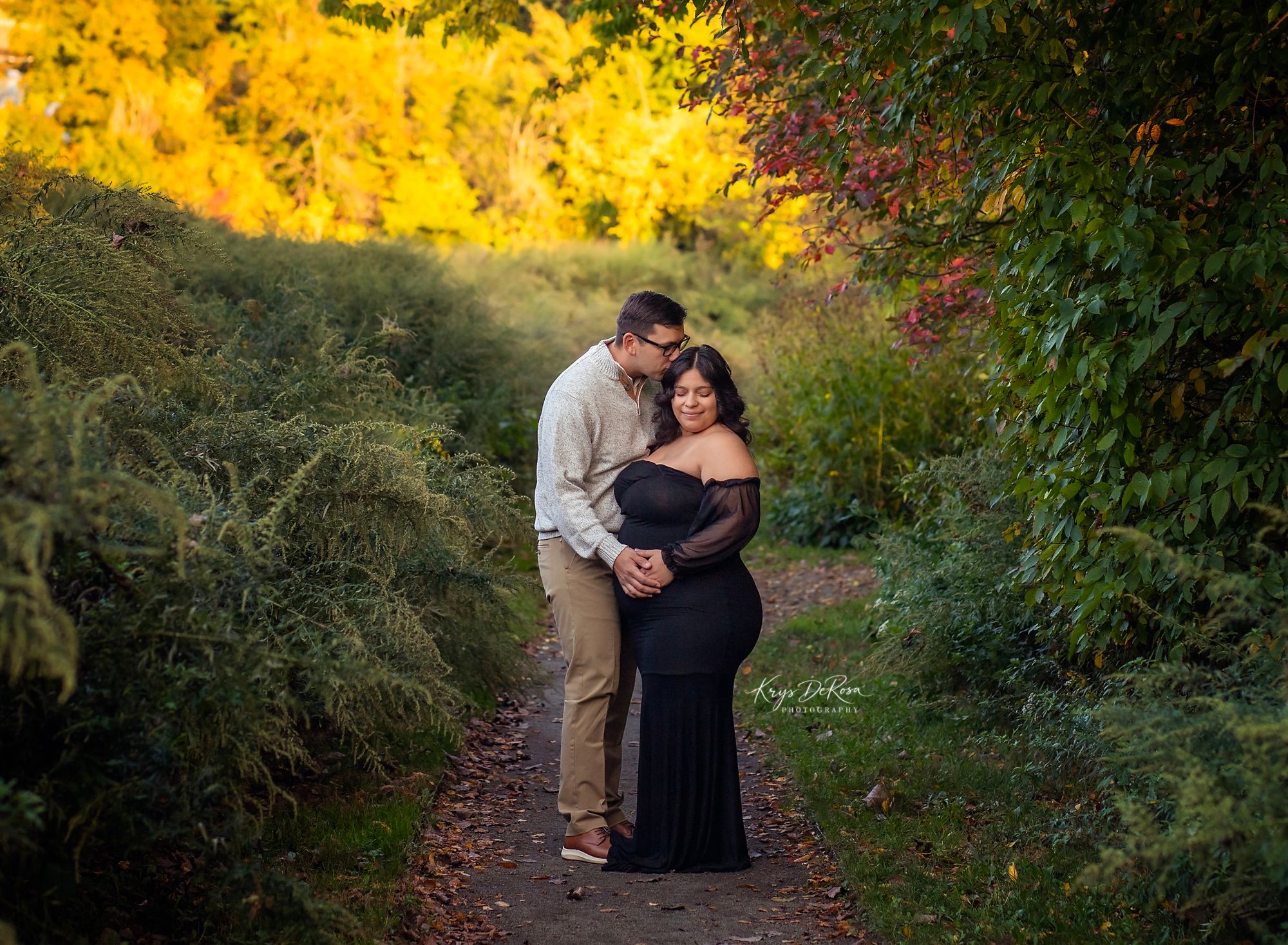 Couple embracing on a garden path surrounded by vibrant autumn foliage during a maternity photo shoot.