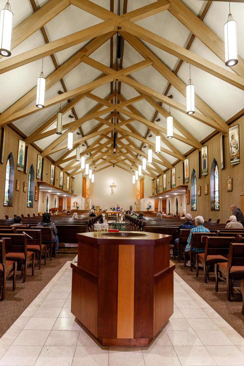 Interior view of a modern church sanctuary with wooden pews and vaulted ceiling featuring pendant lights and stained glass windows.