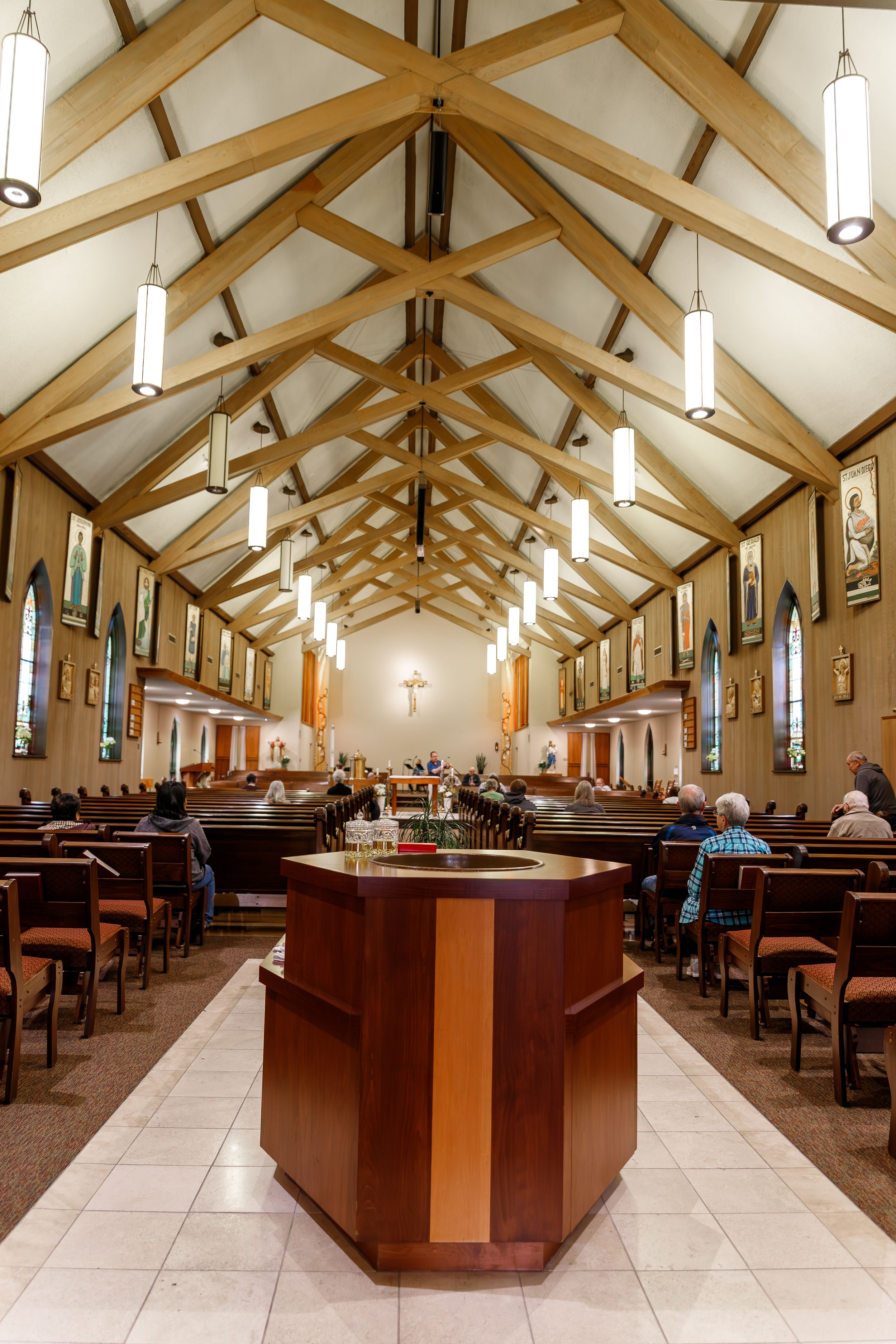 Interior view of a modern church sanctuary with wooden pews and vaulted ceiling featuring pendant lights and stained glass windows.