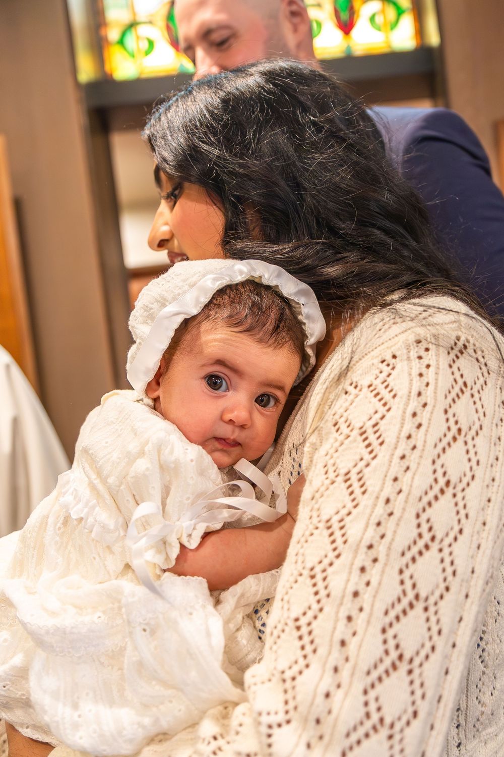 At a baptism ceremony, a baby in white christening gown is held closely in white lacy clothing.