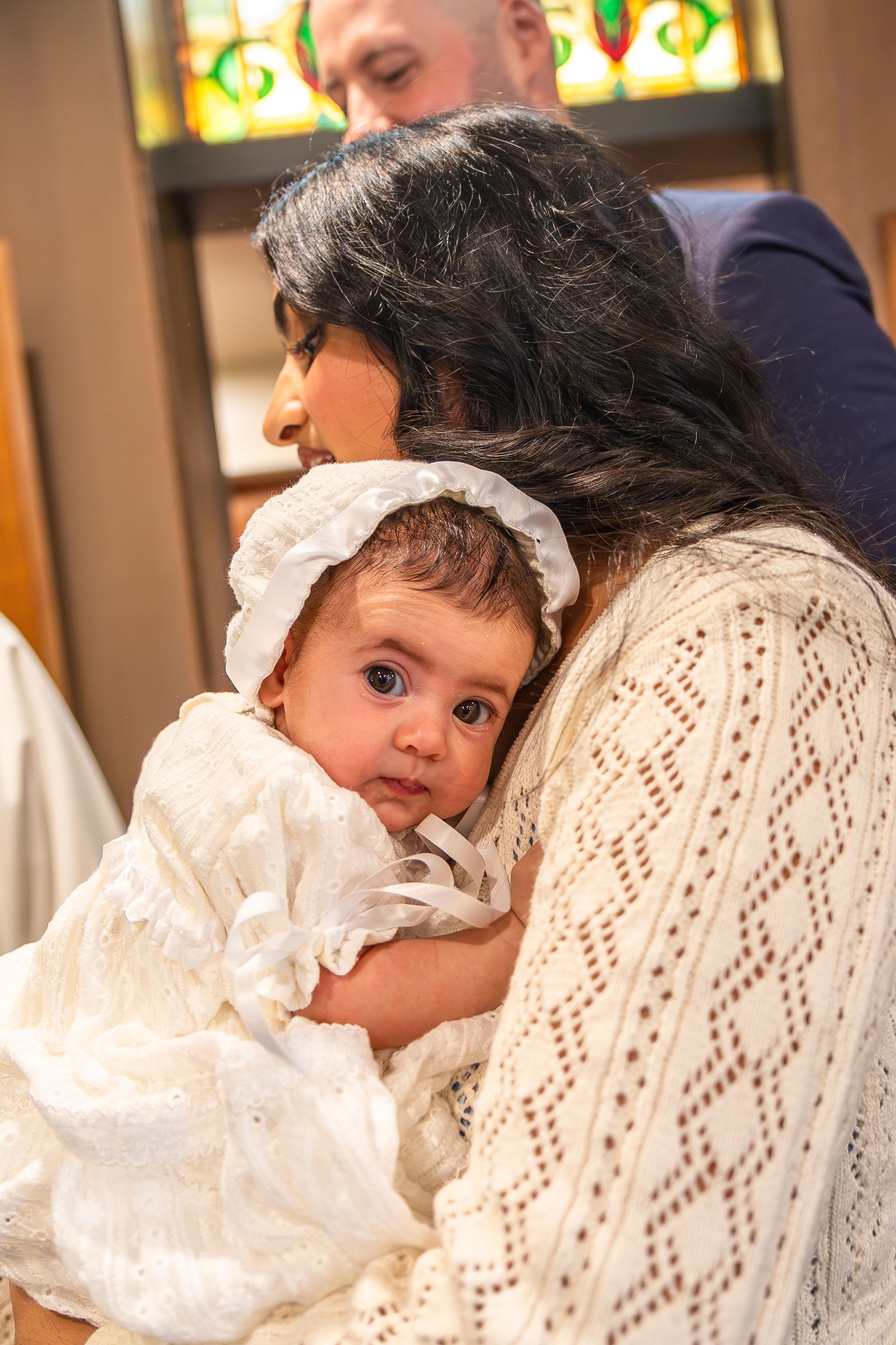 At a baptism ceremony, a baby in white christening gown is held closely in white lacy clothing.