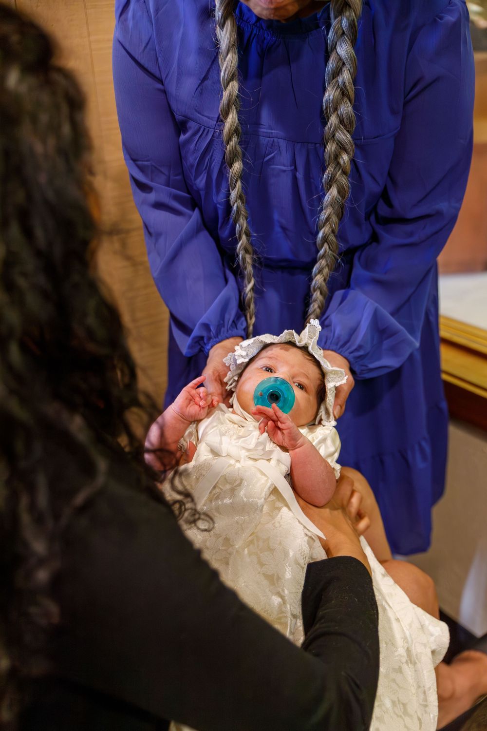 Baby wearing white christening gown being held by someone in a blue outfit during a baptism ceremony.