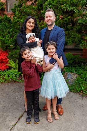 A family of five poses together in front of lush greenery with bright red flowers.