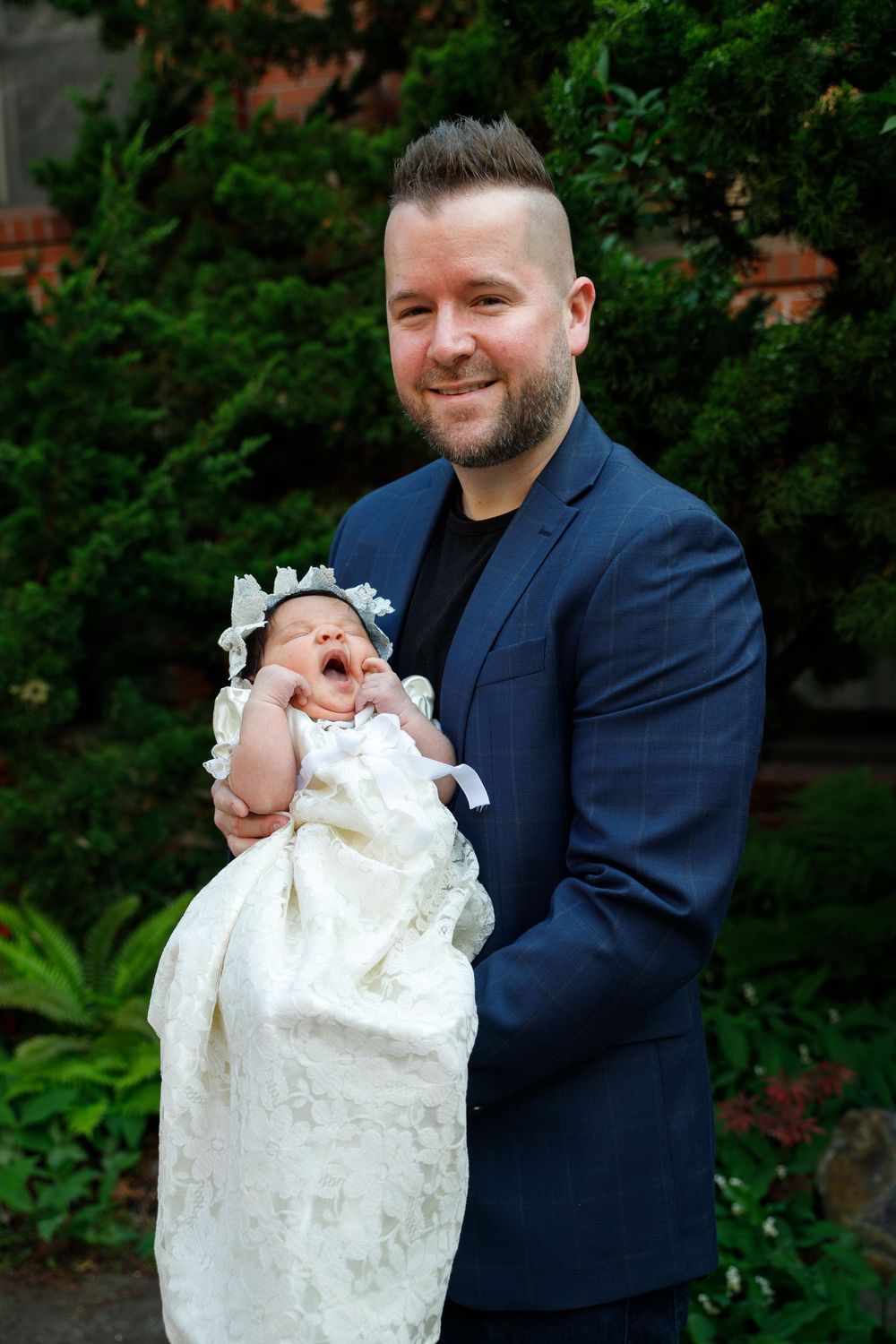 Baby in an ornate white christening gown being held outdoors in a garden setting.