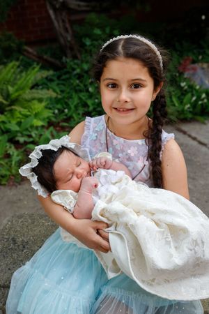 A young girl in a blue dress holds a sleeping newborn baby wearing a white christening gown in a garden setting.