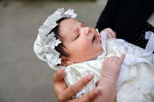 A sleeping infant in a white christening gown and lace bonnet rests in gentle embrace.
