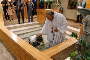 A priest in white robes performs a baptism ceremony in a wooden baptismal font.