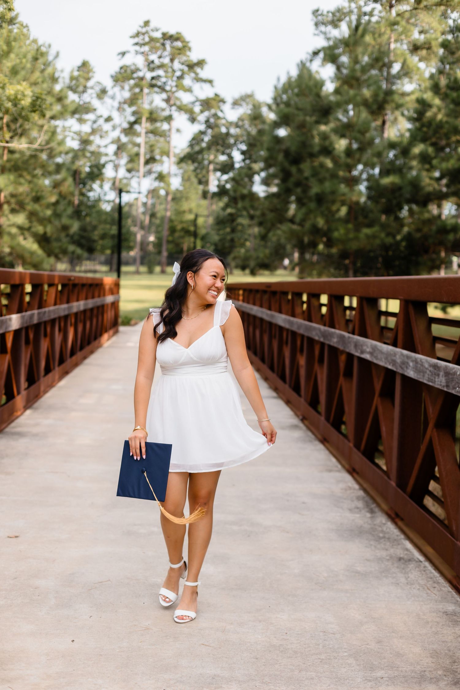Smiling young woman in a white dress holding a graduation cap while walking on a wooden bridge.