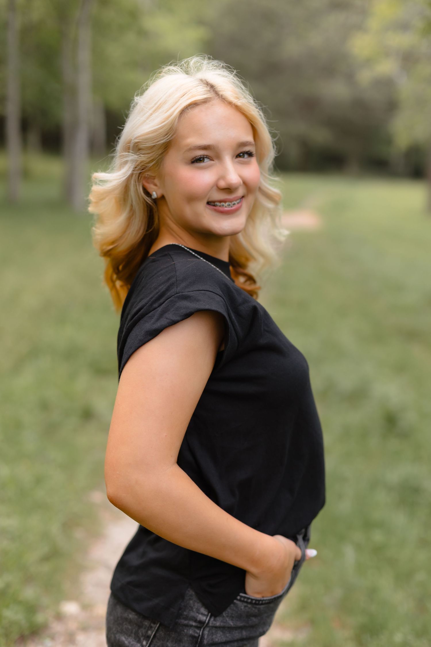 Smiling blonde teenage girl with braces in black t-shirt posing outdoors in green park for senior portrait.