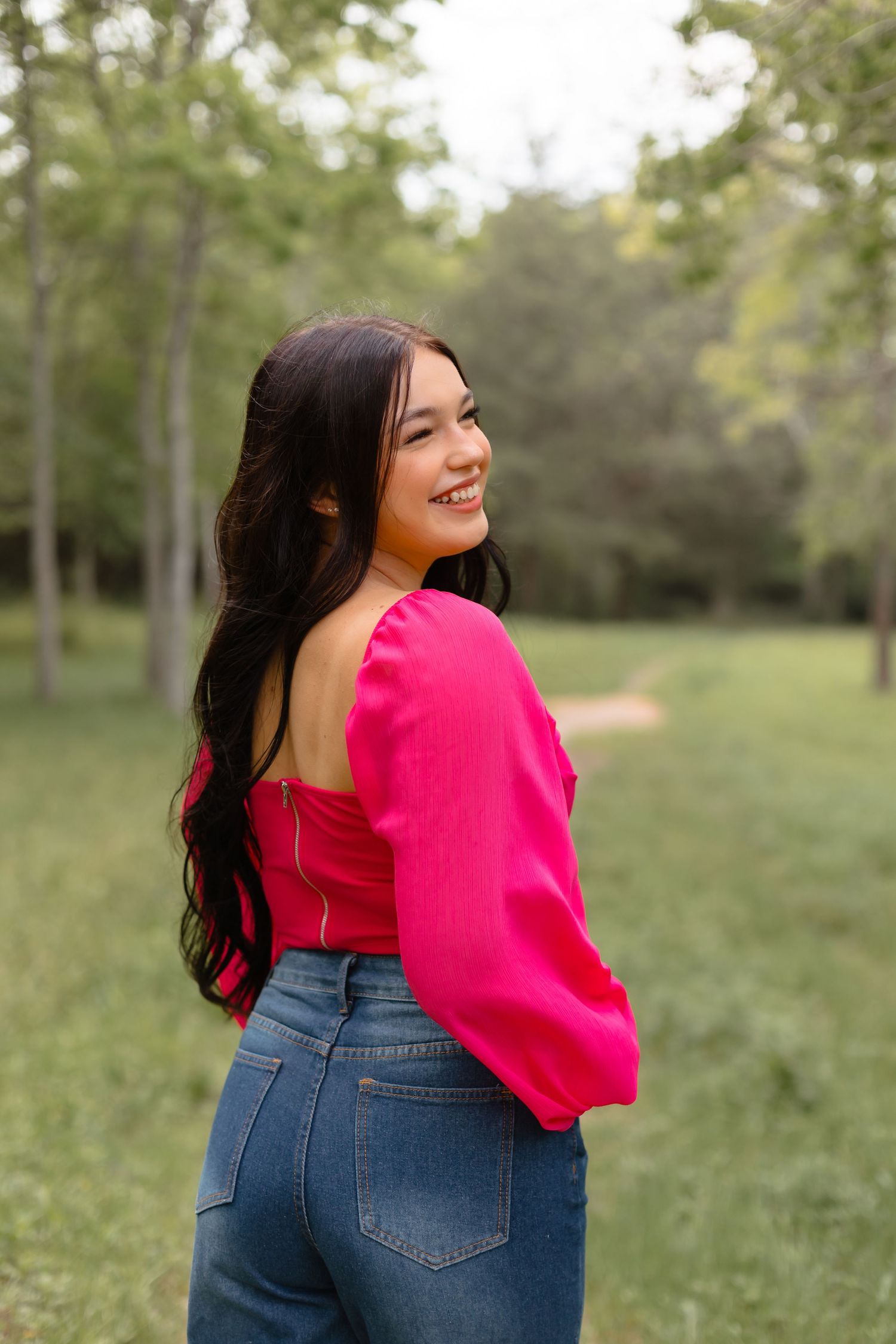 Brunette young woman in pink off-shoulder top and jeans smiling over shoulder in outdoor park setting.
